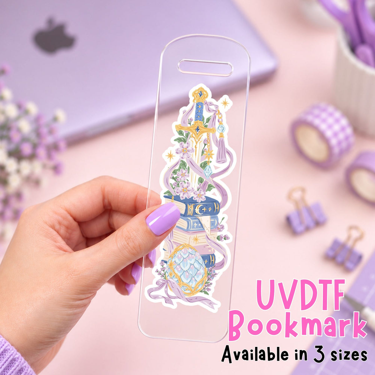 A hand with lavender nail polish holds a clear acrylic bookmark decorated with a colorful fantasy book and sword illustration. The background is a soft pink desk with a purple laptop, washi tape, binder clips, and other stationery items.
