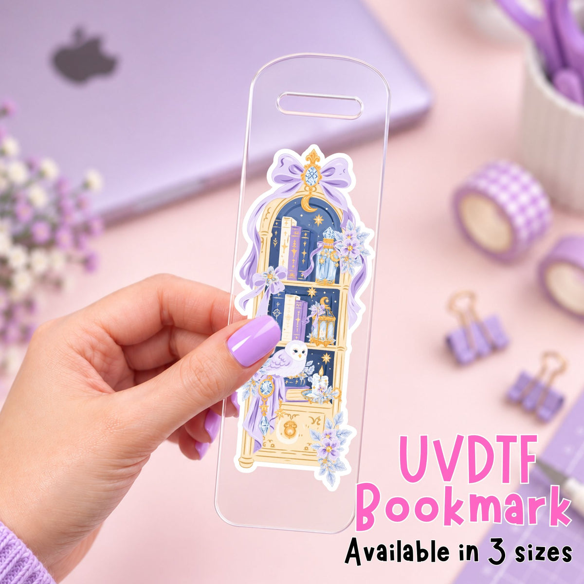 A hand holds a clear acrylic UVDTF bookmark featuring a detailed illustration of a purple and gold bookshelf with books, flowers, and bows. The background is a soft pink desk setup with a laptop, washi tape, binder clips, and other stationery items.