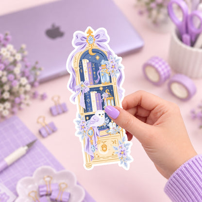 A hand with lavender-painted nails holds a decorative sticker featuring a purple and gold bookshelf illustration with flowers. The background is a styled desk setup in pastel purple tones with a laptop, washi tape, clips, and flowers.