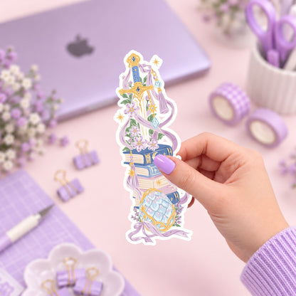 A hand with lavender nail polish holds a die-cut sticker featuring a fantasy illustration of a golden sword, books, flowers, and a mermaid tail. The background is a pastel desk setup with a purple laptop, washi tape, and stationery.