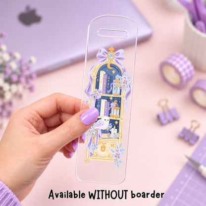 A hand with lavender nail polish holds a clear phone case featuring a detailed illustration of a purple and gold bookshelf with books, flowers, and a cat. The background is a soft pink desk with a purple laptop, washi tape, and paper clips, with text at the bottom reading 'Available WITHOUT boarder'.