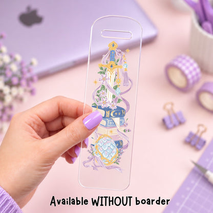 A hand with lavender nail polish holds a clear phone case featuring a detailed illustration of a fantasy book tower with purple ribbons, flowers, and gold accents. The background is a soft pink desk with purple-themed stationery, including a laptop, washi tape, and binder clips.