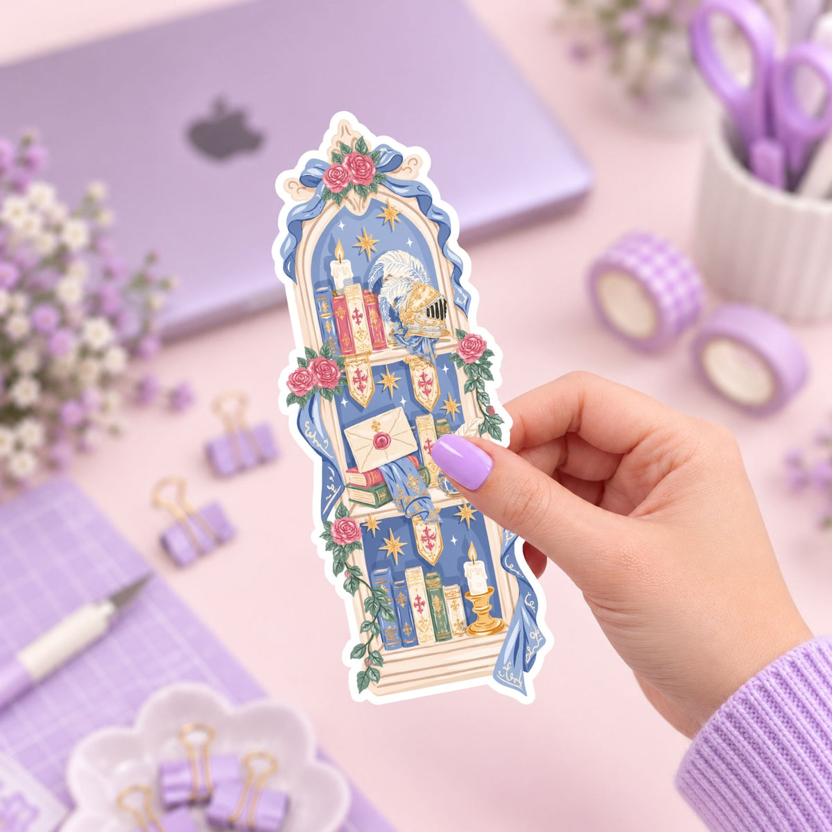 A hand with lavender-painted nails holds a die-cut sticker featuring a whimsical library illustration with books, candles, stars, and flowers. The background is a styled desk setup in pastel purple and pink tones with a laptop, washi tape, scissors, and clips.