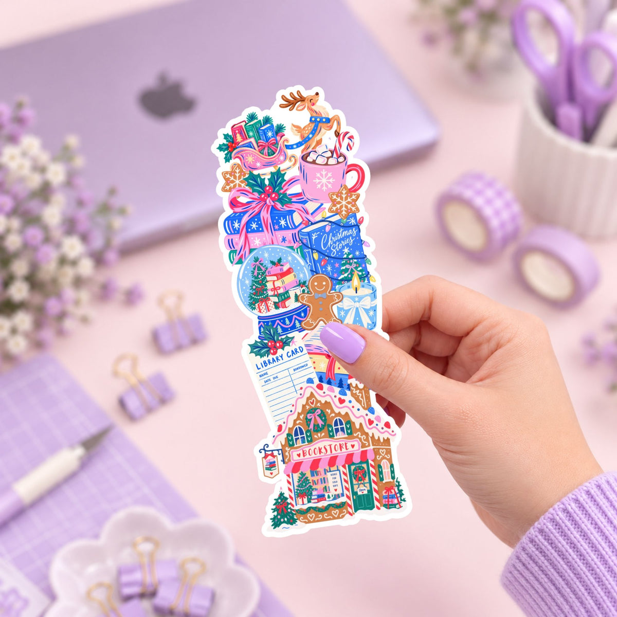 A hand with lavender-painted nails holds a colorful Christmas-themed sticker sheet featuring a gingerbread house, gifts, snow globe, mug, and holiday decorations, set against a pastel purple desk background with a laptop, washi tape, and stationery.