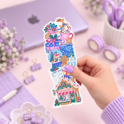 A hand with lavender-painted nails holds a colorful Christmas-themed sticker sheet featuring a gingerbread house, gifts, snow globe, mug, and holiday decorations, set against a pastel purple desk background with a laptop, washi tape, and stationery.