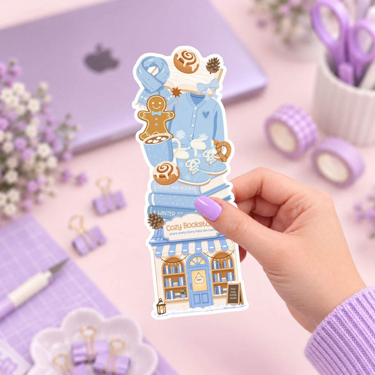 A hand with lavender nail polish holds a die-cut sticker featuring a cozy blue bookshop and gingerbread cookies, set against a pastel purple desk background with stationery items.