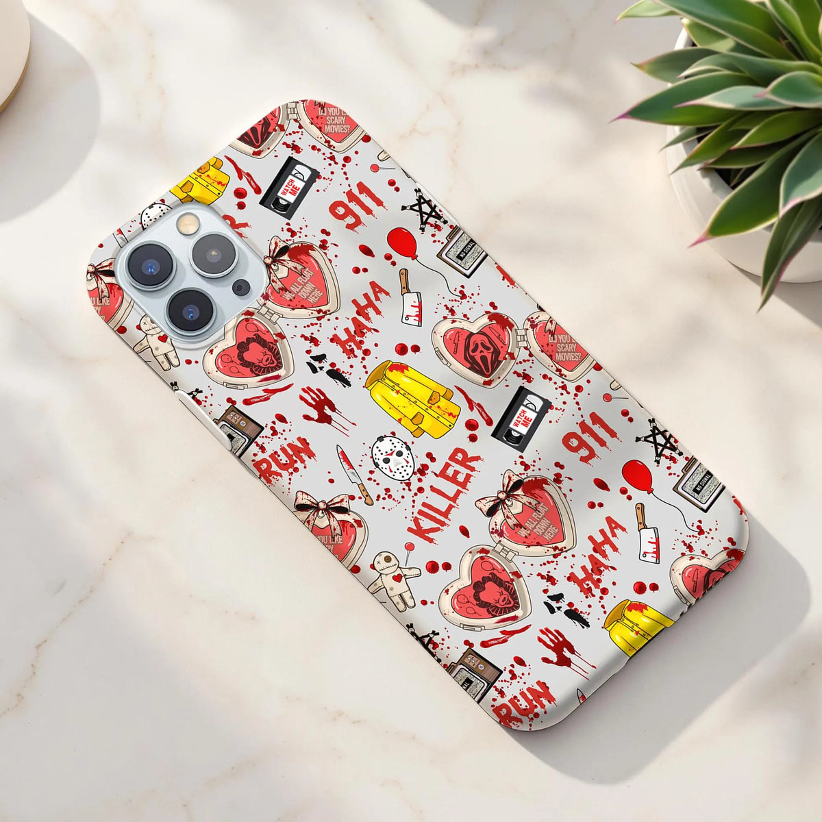 A phone case with a white background and a red, black, and yellow graphic pattern featuring hearts, cassette tapes, blood splatters, and text like 'BATH', 'KILLER', and '911', shown on a marble surface next to a plant.
