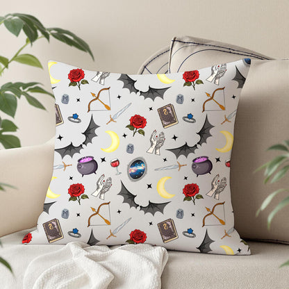 A square throw pillow with a whimsical romantasy-themed pattern featuring dragons, cauldrons, roses, and stars on a white background, styled on a beige sofa with house plants.