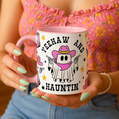 Person holding a mug with a cartoon ghost in a cowboy hat and boots design, text 'YEEHAW AND HAUTIN', and pink handle.